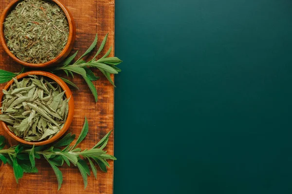 Stevia kuru yapraklar ve yuvarlak ahşap kaselerde toz ve koyu yeşil arka planda tahta üzerinde taze bitki dalları.