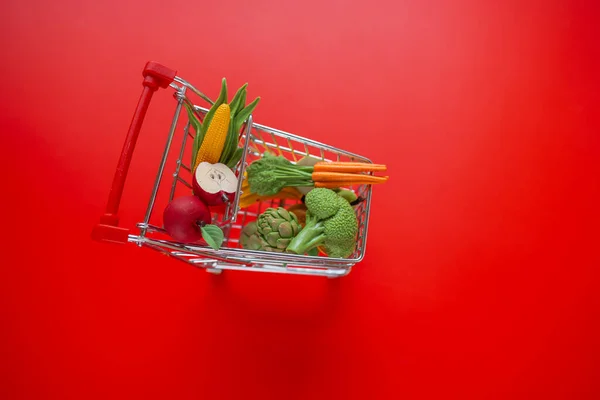 Shopping cart with groceries on a red background.grocery consumer basket.food cost.Vegetables and fruits price increase.Decorative supermarket trolley with groceries on a red background.