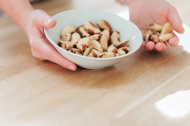 Fındık ve tohum. Brezilya fındığı elinde beyaz bir seramik bardakta ahşap bir masada. Diyetteki sağlıklı yağlar. El, Brezilya fındıklarını yakın plandan alıyor. Faydalı sağlıklı atıştırmalıklar..