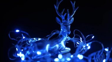 Karanlık bir arka planda Noel geyiği ve çelenk. Karanlıkta yanıp sönen mavi ışıktaki gümüş geyik. 4k görüntü