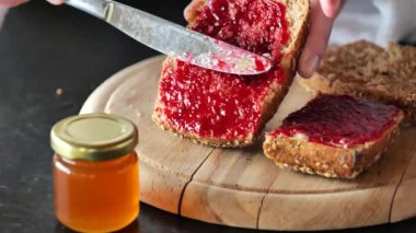 Ahududu reçelli tam tahıllı çörek. Kahvaltı için geleneksel yemek. Kadınların elleri bütün bir tahıllı çöreğe ahududu reçeli sürer. Kahvaltıyı hazırlar. 4k görüntü