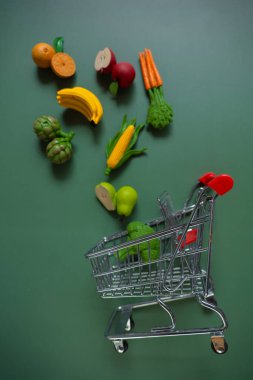 Alışveriş yemeği. Yeşil zemin üzerinde dekoratif sebzeler ve meyvelerle süpermarket arabası. Artan gıda fiyatları. Sebze ve meyve fiyatları artışları.