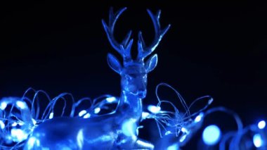 Noel arkaplanı. Karanlık bir arka planda geyik ve çelenk. Karanlıkta yanıp sönen mavi ışıktaki gümüş geyik. 4k görüntü