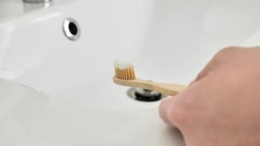 Banyoda musluk suyunun altında elinde diş macunu olan bambu diş fırçası. Diş temizliği. El, parlak bir banyoda diş fırçasını yakın plan çeker. Yavaş hareket. İnsan hijyeni konsepti. 4k görüntü