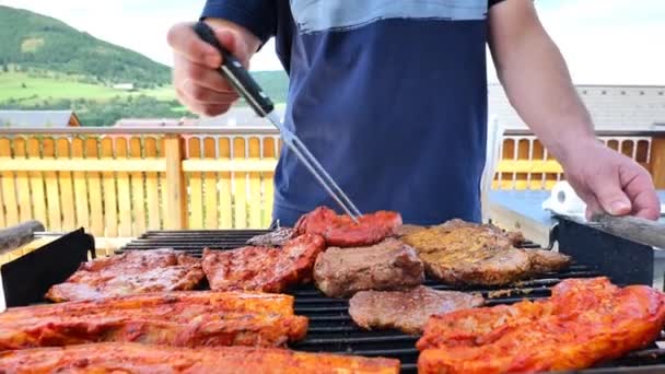 Bbqとグリル 青いTシャツの男は 長いフォークで焼いた肉を回します 肉はグリルの上に回ります 揚げた豚肉 焼肉4K映像付き屋外ピクニック — ストック動画