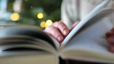 Noel kitapları. Elleri açık renk bir kazak içinde, arka planda çelenkleri olan bir Noel ağacının sayfalarını çevirin. Noel ve kış temalarıyla ilgili kitaplar. Kış kitapları..