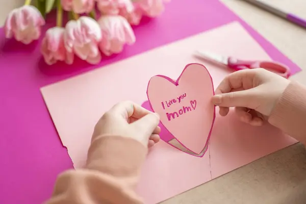 Mothers Day.I love you, Mom.Flowers and cards for mom.Childrens hands holding a heart card.Pink heart card and pink tulips.child makes a card for his mother.moms day concept.
