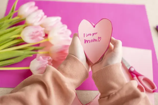 Mothers Day.I love you, Mom. Childrens hands holding a heart card.Pink heart card and pink tulips.child makes a card for his mother.Flowers and cards for mom.moms day concept.