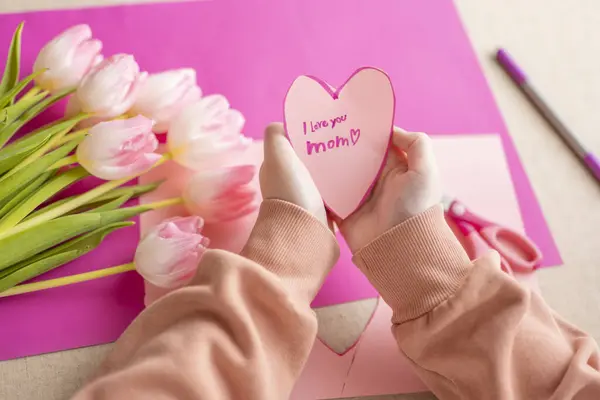 I love you, Mom. Childrens hands holding a heart card.Pink heart card and pink tulips.child makes a card for his mother.Flowers and cards for mom.moms day concept.