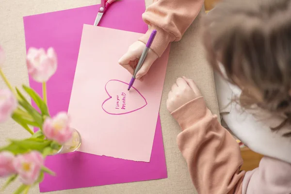 Mothers Day.Flowers and cards for mom.Daughter draws a card for mom.Message to mom.Child draws a heart on a pink piece of paper and writes mom I love you.