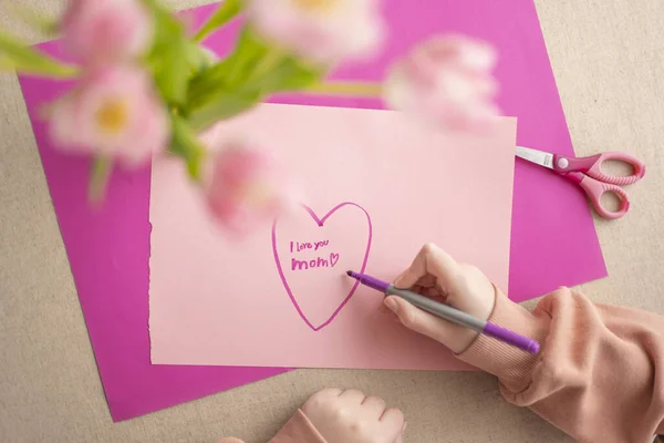 moms day.DIY mom card.Mothers Day.Child draws a heart on a pink piece of paper and writes mom I love you. daughter makes a card for his mother.Flowers and cards for mom.Daughter draws a card for mom.