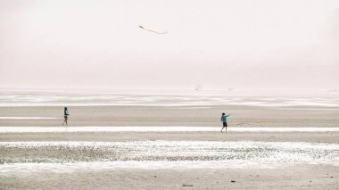 Uçurtmalar ve insanlar beyaz kumlu deniz sahillerinde yürüyorlar. Deniz kıyısında uçurtma uçuruyorlar. Deniz kenarında tatil yapıyorlar. İnsanlar bulutlu bir günde uçurtma uçuruyorlar..