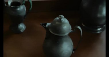 Çaydanlıklar, tahta bir rafta teneke sürahiler, teneke çanak ve teneke çatal bıçak takımı. Cam bir vitrinde metal antikalar. Klasik sofra takımı. 4k görüntü