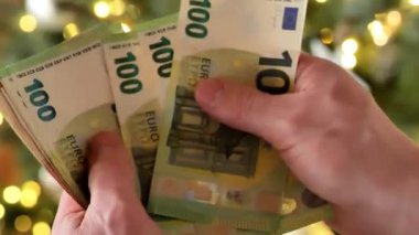 Noel harcamaları ve harcamaları. Tatil masrafları. Noel ve yeni yıl alışverişleri ve alışverişleri. Erkek eller, arka planda çelenkler olan bir Noel ağacında euro banknotlarını sayıyor. 4k görüntü