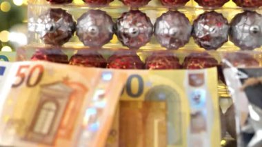 Tatil harcamaları. Avro banknotları ve Noel ağacı arkaplanındaki parlak çelenklerin üzerindeki kutularda Noel oyuncakları. Noel süslemeleri ve hediyeleri almak. 4k görüntü