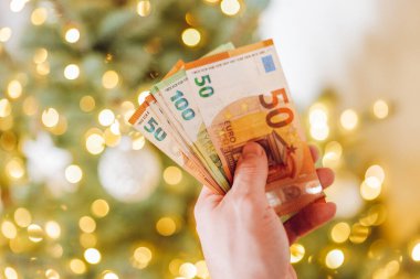 Euro banknotları ve Noel ağacı. Noel Tatili Masrafları. Noel ve Yeni Yıl Alışveriş ve Alışverişleri. Yılbaşı ağacındaki euro banknotlarını sayan eller. Tatil masrafları ve harcamalar..