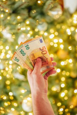 Noel Tatili Masrafları. Euro banknotları ve Noel ağacı. Noel ve yeni yıl alışverişi. Yılbaşı ağacındaki avro banknotlarını sayan eller..