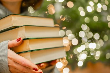 Kış tatillerinde Noel okuması. Noel ağacının arka planında yeşil kapaklı kitap yığınını tutan eller. Sıcak kış tatili havası..