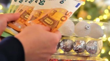 Noel süslemeleri alıyorum. Altın Bokeh çelenklerindeki euro banknotlarını sayıyorum. Para, Noel oyuncakları ve süslemeler. Geleneksel Noel Alışverişi. Kış tatili atmosferi. 4k
