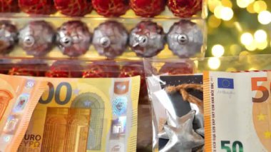 Para, Noel oyuncakları ve dekorasyonlar. Tatil masrafları. Altın çelenkler üzerine Avro banknotları ve parlak Noel ağacı süslemeleri. Noel harcamaları ve harcamaları. 4k görüntü