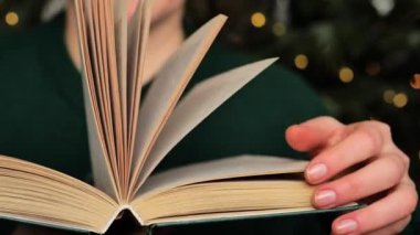 Noel okumaları. Yeşil kazaklı bir kadın, Noel ağacında yeşil bir çelenk takıp kitabı karıştırıyor. Yılbaşı hikayeleri, efsaneler ve efsaneler.