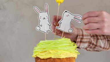 Paskalya pastası için kağıt tavşanlar ve çiçekler. Kağıt Başlıklı Paskalya Fırını ve Yenilebilir Şeker Çimi. Kareli gömlekli eller, paskalya pastasını kağıt çalı ağacıyla süslüyor.. 