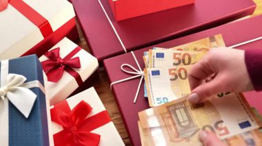 Hediye alışverişi. El, hediye kutularının yanında para sayar. Tatil Hediyesi Parası. Kurdeleli paketlenmiş hediyeler ve Avro banknotları bir masanın üzerinde. Paketlenmiş hediye kutuları üzerinden para sayan eller. 4k görüntü