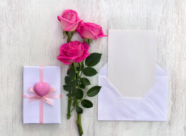 Pink flowers roses, postal envelope with paper card note with space for text, gift box with pink heart on a white wooden background. Top view, flat lay
