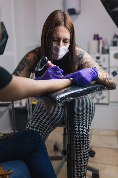 Tattoo artist tattooing in her studio with gloves and mask. Tattoo arm.