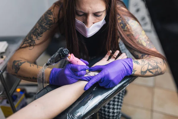 Tattoo artist tattooing in her studio with gloves and mask. Tattoo arm.