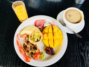 Domuz pastırması, jambon, et, mango, meyve, kahve fincanı ve meyve suyu olan bir masada besleyici ve ferahlatıcı bir yemek manzarası..
