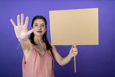Bir elinde tabela tutan genç bir kadın ve mor izole edilmiş arka planda dur işareti yapıyor.