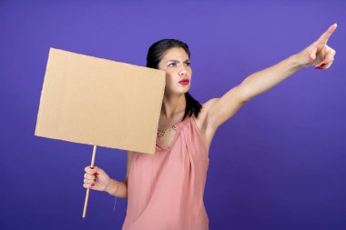 Genç yetişkin bir kadın, bir eli yukarıda, izole edilmiş mor arka planı işaret eden bir tabela tutuyor.