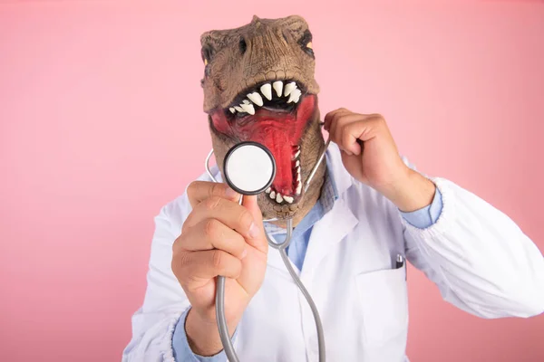 T-Rex maskeli bir doktor, izole edilmiş pembe arka planda steteskop tutuyor. seçici odak