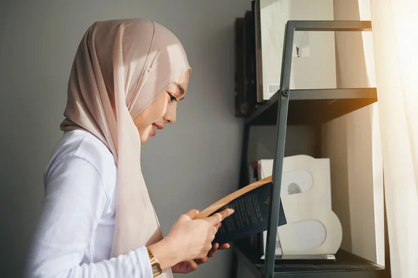 Woman wearing an Islamic hijab headscarf stands reading a book by a shelf near the window.