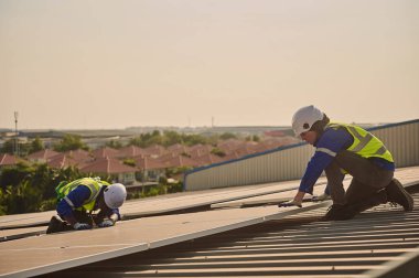 Farklı uluslardan iki mühendis yüksek binalarda çalışırken koruyucu kıyafetler giyerek bir binanın çatısına güneş panelleri kuruyor..