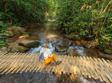 Genç bir kadın Tayland ormanında küçük bir bambu köprüsünde yürüyor.
