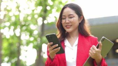 Asyalı iş kadını cep telefonu kullanıyor, akıllı telefonuyla modern ofis dışındaki iş arkadaşlarıyla mesajlaşıyor ve sohbet ediyor..
