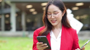 Asyalı iş kadını cep telefonu kullanıyor, akıllı telefonuyla modern ofis dışındaki iş arkadaşlarıyla mesajlaşıyor ve sohbet ediyor..