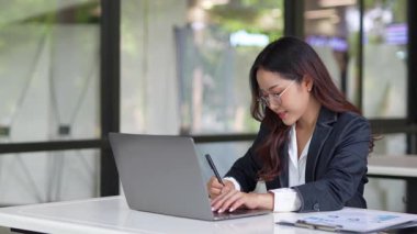 Masada çalışan Asyalı iş kadını dizüstü bilgisayar ve not defteri kullanarak müşteri ya da ofis binasıyla konuşuyor, yavaş çekim görüntüleri