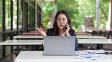 Masada çalışan Asyalı iş kadını dizüstü bilgisayar ve not defteri kullanarak müşteri ya da ofis binasıyla konuşuyor, yavaş çekim görüntüleri