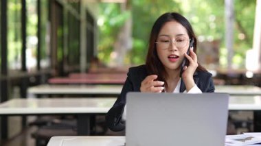 Masada çalışan Asyalı iş kadını dizüstü bilgisayar ve not defteri kullanarak müşteri ya da ofis binasıyla konuşuyor, yavaş çekim görüntüleri