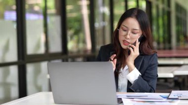 Masada çalışan Asyalı iş kadını dizüstü bilgisayar ve not defteri kullanarak müşteri ya da ofis binasıyla konuşuyor, yavaş çekim görüntüleri