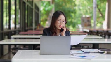 Masada çalışan Asyalı iş kadını dizüstü bilgisayar ve not defteri kullanarak müşteri ya da ofis binasıyla konuşuyor, yavaş çekim görüntüleri