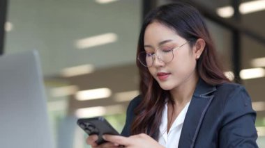 Asyalı iş kadını cep telefonu kullanıyor, akıllı telefonuyla modern ofis dışındaki iş arkadaşlarıyla mesajlaşıyor ve sohbet ediyor..