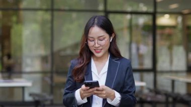 Asyalı iş kadını cep telefonu kullanıyor, akıllı telefonuyla modern ofis dışındaki iş arkadaşlarıyla mesajlaşıyor ve sohbet ediyor..