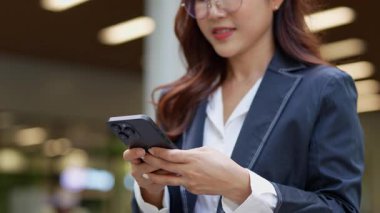 Asyalı iş kadını cep telefonu kullanıyor, akıllı telefonuyla modern ofis dışındaki iş arkadaşlarıyla mesajlaşıyor ve sohbet ediyor..
