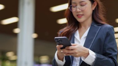 Asyalı iş kadını cep telefonu kullanıyor, akıllı telefonuyla modern ofis dışındaki iş arkadaşlarıyla mesajlaşıyor ve sohbet ediyor..