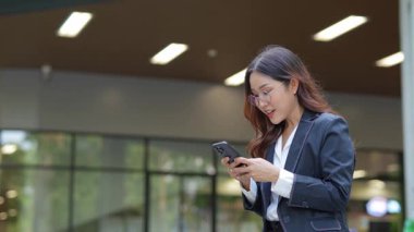 Asyalı iş kadını cep telefonu kullanıyor, akıllı telefonuyla modern ofis dışındaki iş arkadaşlarıyla mesajlaşıyor ve sohbet ediyor..