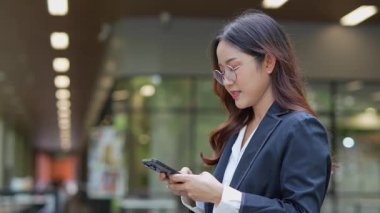Asyalı iş kadını cep telefonu kullanıyor, akıllı telefonuyla modern ofis dışındaki iş arkadaşlarıyla mesajlaşıyor ve sohbet ediyor..
